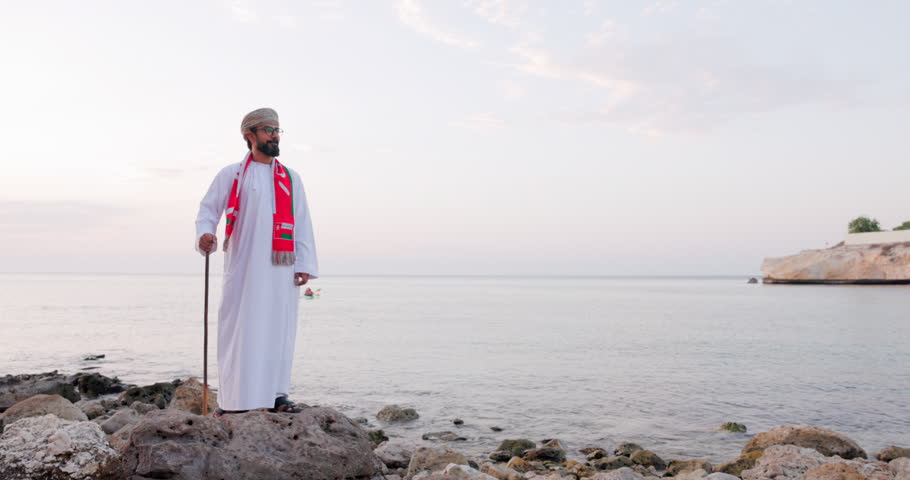 Omani man in a red scarf around his neck and traditional walking stick stands on a rocky shoreline