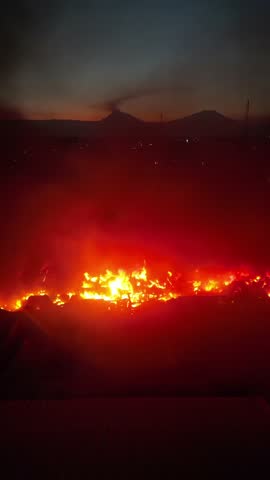 Flames engulf a factory, sending thick black smoke billowing into the evening sky. The intense fire contrasts with the darkening surroundings, creating a dramatic and alarming scene.