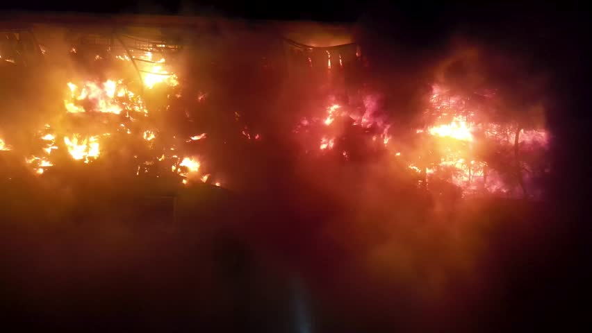 Flames engulf a factory, sending thick black smoke billowing into the evening sky. The intense fire contrasts with the darkening surroundings, creating a dramatic and alarming scene.