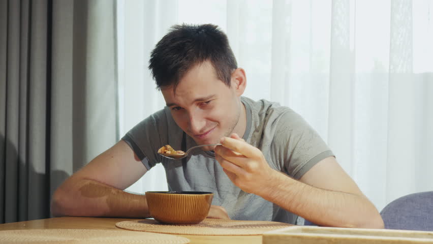 A young man sitting at the dining table, grimacing as he looks at his spoon, struggling to eat a dish he dislikes.