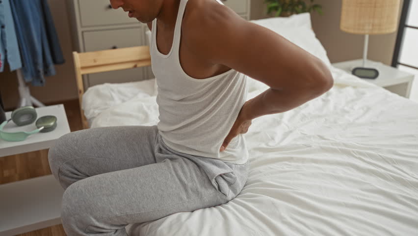 Young man in white tank top and gray pants sitting on bed in bedroom experiencing back pain, with bedside table, flowers, and clothing visible in the interior.