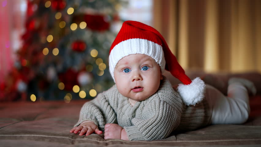 Adorable Caucasian baby with big blue eyes lies looking at camera. Lovely baby boy wearing Santa cap and warm sweater. Blurred Christmas tree at backdrop.