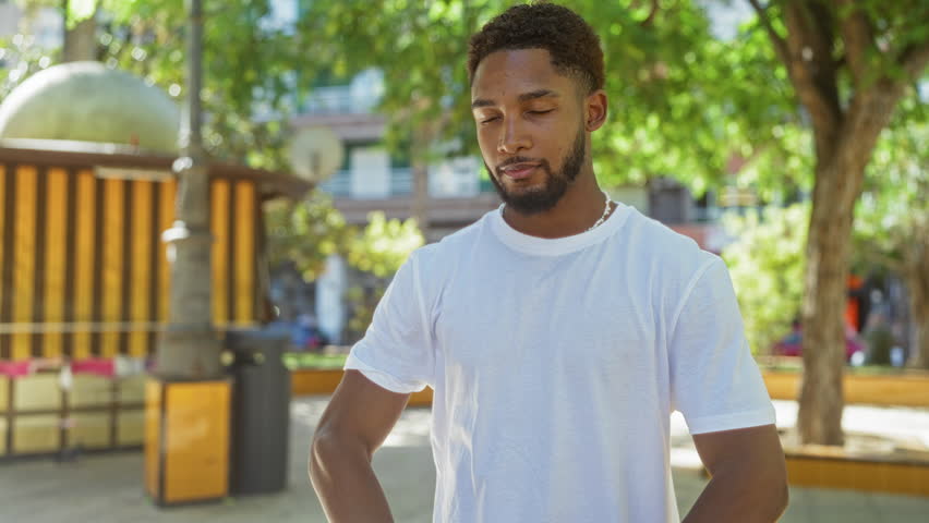 Young man meditating outdoors in an urban park, with a serene expression and trees providing shade around the cityscape background
