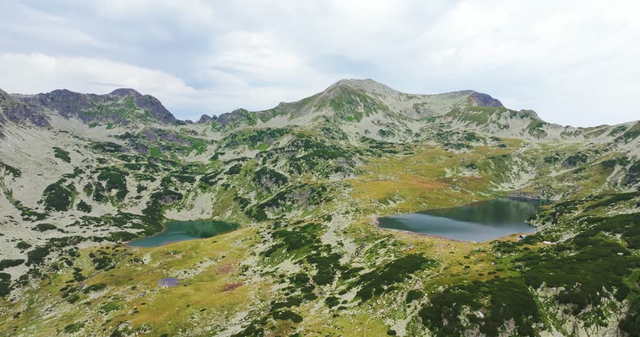 Multiple glacial lakes spread across vibrant green pastures surrounded by tall mountain ranges, rising drone shot