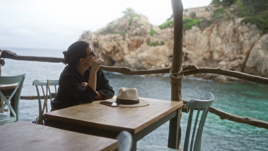 Young hispanic woman sitting outdoors at a seaside cafe in cala deia, mallorca, spain, with hat and smartphone on a wooden table, enjoying ocean view and relaxed atmosphere