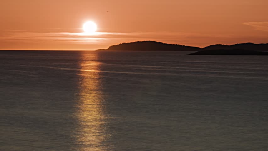 A beautiful sunset paints the sky orange as it dips behind distant islands over calm Norwegian waters.