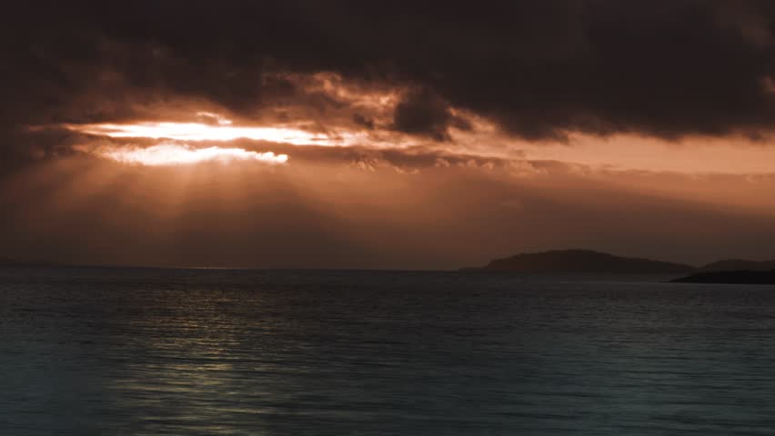 A dramatic sunset over the calm sea, with the horizon broken by dark, distant islands in the evening light.