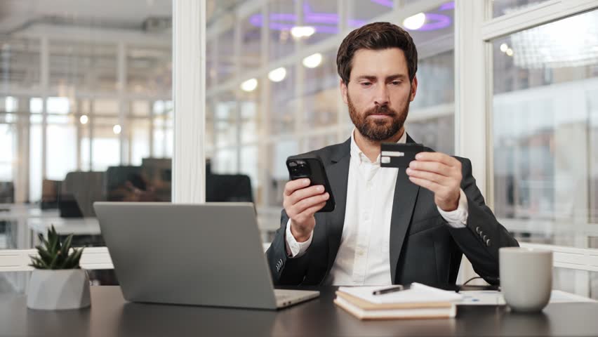 Confused businessman in a suit sits at his desk in an office, angry millennial male hold prepaid card look on mobile feel mad annoyed get payment rejection after entering wrong password