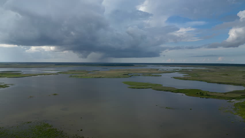 Aerial view of a vast wetland landscape under dramatic storm clouds in Semigallia, showcasing large water bodies surrounded by lush green marshes, with a rainstorm approaching in the distance. 4K.