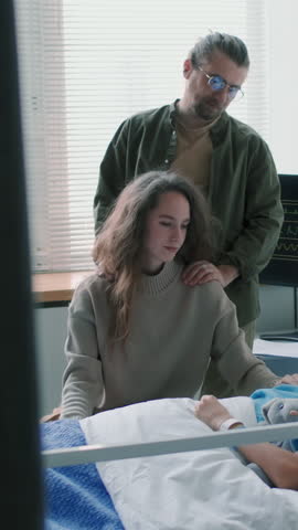 Vertical shot of young Caucasian parents staying beside sleeping sick child who lying in hospital bed during complication of disease period in ward decorated with balloons