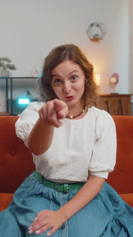 Hey you. Young woman smiling excitedly and pointing to camera, choosing lucky winner, indicating to awesome you, inviting, approve. Portrait of girl at home living room sitting on couch. Vertical.