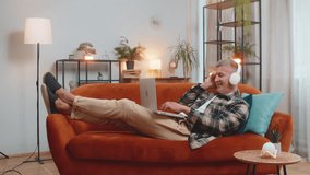 Happy relaxed Caucasian young man with gray hair in wireless headphones lying on home couch sofa choosing listening disco music while working on laptop netbook. Guy weekends activities entertaining - Powered by Shutterstock - Get 15% off with code: PIKWIZARD15