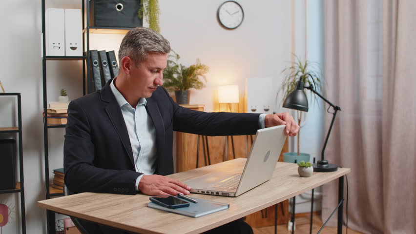 Young gray-haired businessman freelancer in formal suit opens laptop start working sends messages, watching movies, makes online purchases. Caucasian guy using netbook in modern home office workspace.