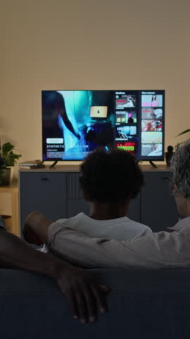 Vertical rear view of diverse family coming to living room to watch movies on streaming service together in evening, active boy jumping onto sofa and parents with popcorn taking seat in front of TV
