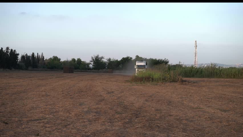 A large truck loaded with hay bales drives on a dirt road in a field, passing the camera