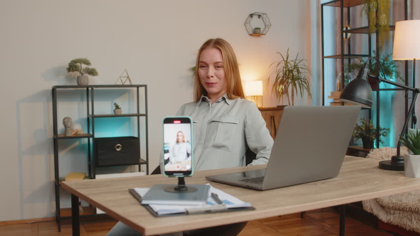 Smiling middle-aged business woman sitting at table before smartphone fixed on tripod record motivation training webinar on web camera. Video blogger influencer broadcast live speech using cell phone.