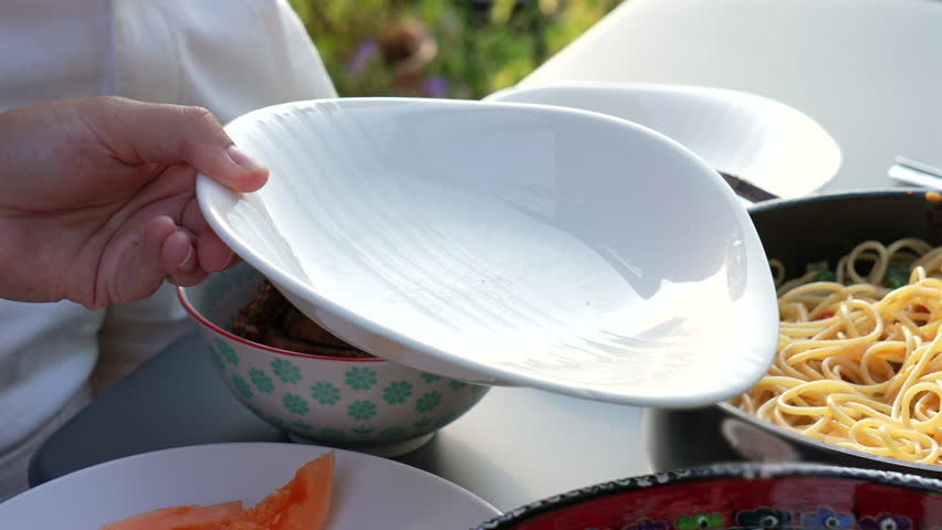 Person serving a portion of spaghetti onto a plate from a pot, with visible pieces of cured meat and basil leaves mixed with the pasta. Outdoor casual dining scene with colorful tableware