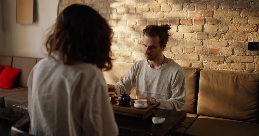 Confident guy with stubble tastes tea and chats with a brunette in a white shirt in a cafe