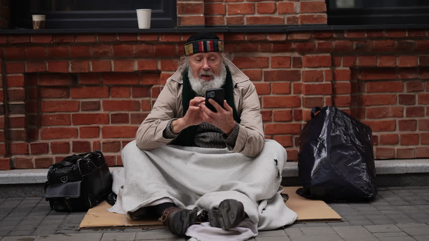 Portrait of tired retired homeless man using smartphone sitting outside on cold day, trying to find shelter or contact someone for help. Older tramp male seek work and government assistance.
