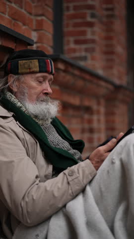 Vertical shot of retired homeless man sits on urban street, wrapped in blanket, using smartphone in front of brick wall, reflecting poverty and social issues. Concept of poor unhappy retirement.