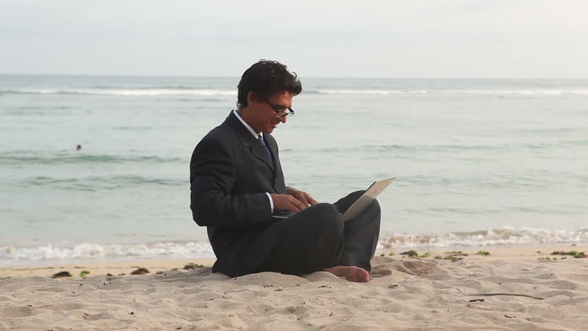 Handsome businessman sitting at beach and typing on laptop