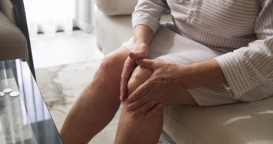 Top view of a pensioner's hands rubbing a popliteal or synovial cyst, a fluid-filled swelling that forms on the back of the knee. Popliteal cyst, fluid in joint. Damaged knee joint of an elderly woman