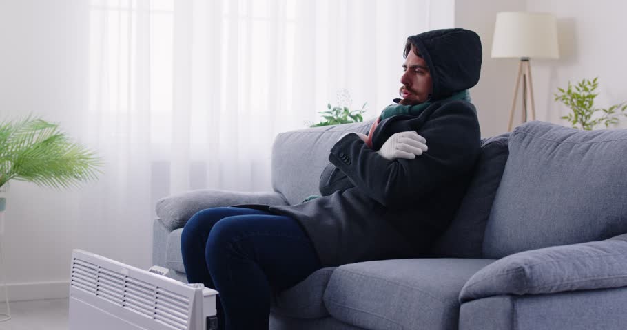 Young man wearing a winter jacket, scarf and gloves sitting on the couch in the living room in his house or apartment, feeling very cold and trying to warm up by the electric heater. Cold home concept