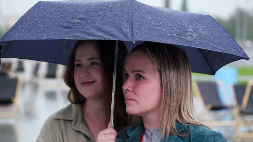Two women joyfully sharing an umbrella while standing together in the rain and sharing a moment. Clip