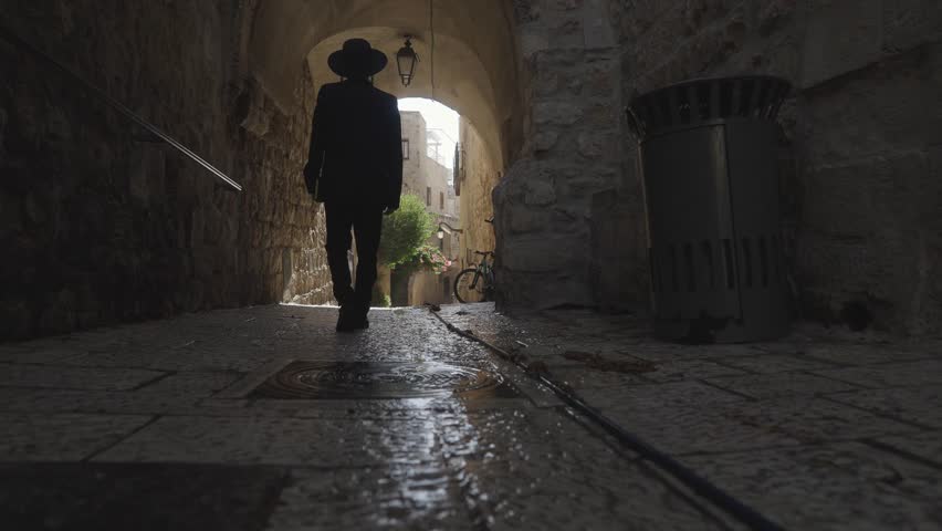 Orthodox Jewish man walking through an ancient passageway in the Old City of Jerusalem, Israel. He has side curls, black hat and suit and holds a prayer book. He is seen from behind in slow motion.