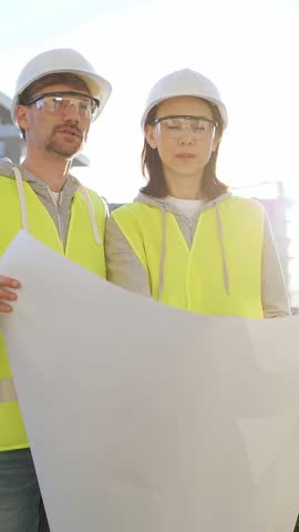 Man and woman architects or engineers wearing safety hard hats and vests holding blueprint and discussing something on construction site at sunrise, front vertical view. Architecture concept