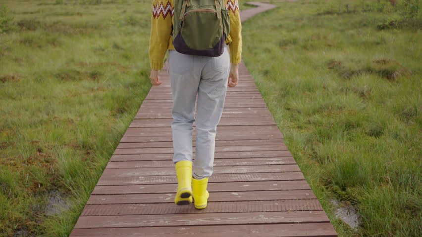 Hiker wearing yellow rain boots walks away on a wooden path, surrounded by lush green grass in a serene nature preserve, enjoying a peaceful moment in the outdoors