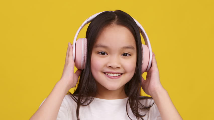 A smiling Asian girl adjusts her pink headphones, enjoying music against a cheerful yellow backdrop.
