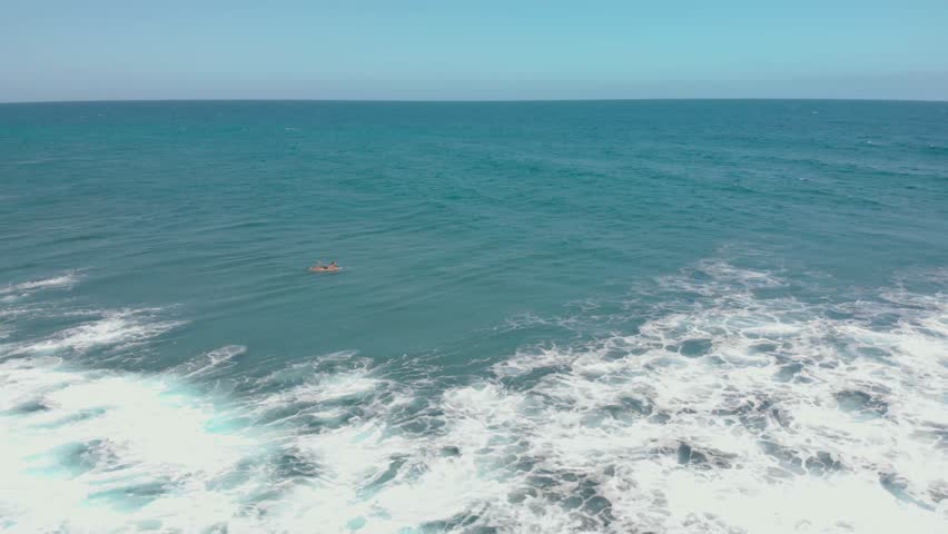 A lone surfer paddles on a surfboard in the vast ocean, surrounded by deep blue and green waves under a clear sky at Aguadilla Beach in Puerto Rico.
