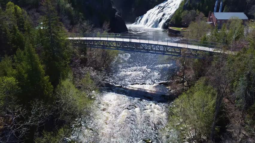 Drone flight under a pedestrian bridge over a river giving a magnificent view of a waterfall from an old hydroelectric power plant. Riviere-du-Loup, Quebec.