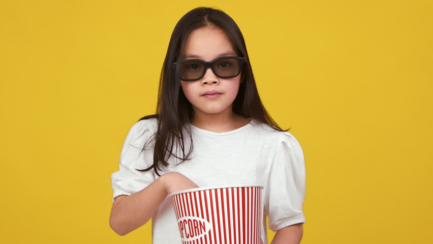 Asian child is munching on popcorn, wearing 3D glasses, with a bright yellow backdrop adding a playful touch.