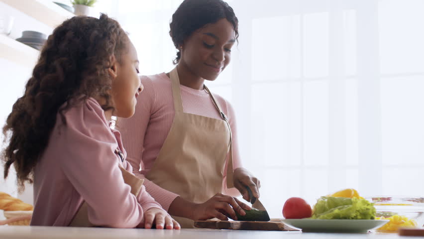 African American mother and daughter enjoy quality time in a well-lit kitchen, preparing fresh vegetables for a healthy meal while sharing smiles and laughter during their cooking session.