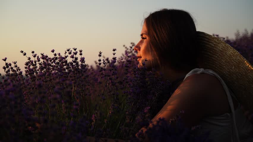 A woman sits in a field of lavender purple flowers. She is wearing a straw hat and hugging lavender flowers.