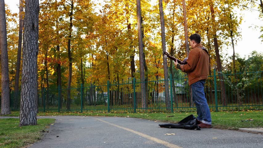 A boy street musician stands on the path in the park, playing guitar and collecting money in his case. The shot is a wide angle.