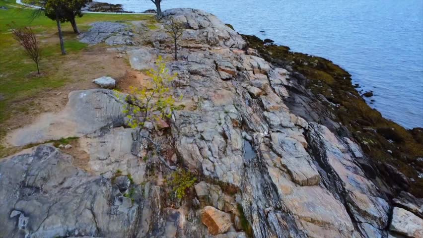 A drone footage of a rocky shoreline during daytime, with green field and coastal trail in the background