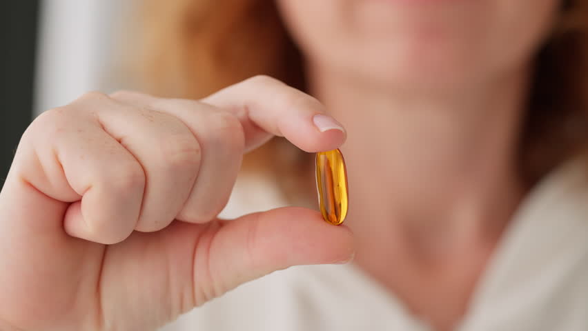 Close-up woman’s hand holding two fingers orange capsule with Omega D3 in camera and puts it near her mouth white background smily. Health, complex vitamins, self-care people. Taking medication, pills