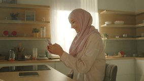 Smiling muslim woman in hijab use phone typing text message chatting in social media sitting at home kitchen at sunlight. Feel happy. Camera moving around. 360 degrees - Powered by Shutterstock - Get 15% off with code: PIKWIZARD15