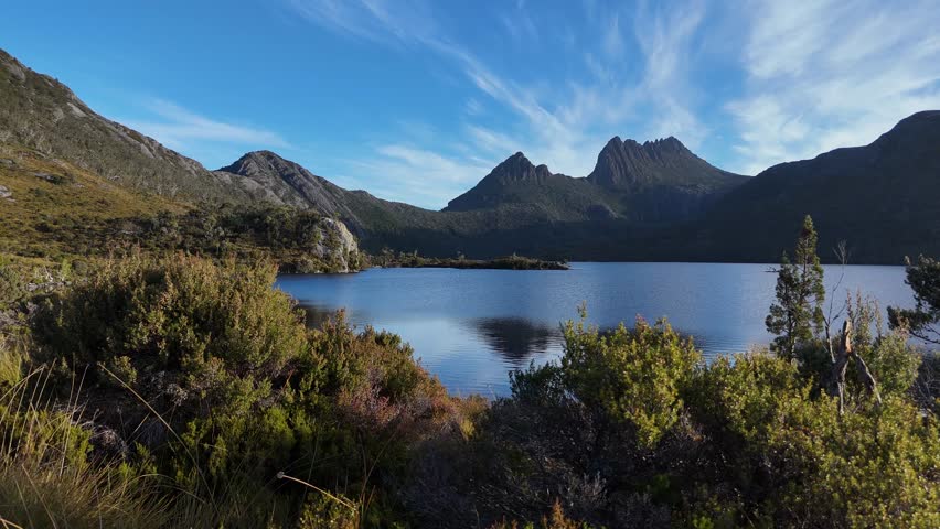 Aerial wide shot of Dove Lake and Cradle Mountain in background. Sunny day with green vegetation in Tasmania, Australia. Wild nature and world heritage area. Low angle lateral shot.