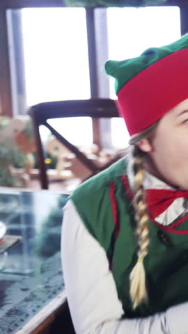 Excited elf sitting at the table. Joyful female in elf green costume is looking at something with surprise. Vertical video.