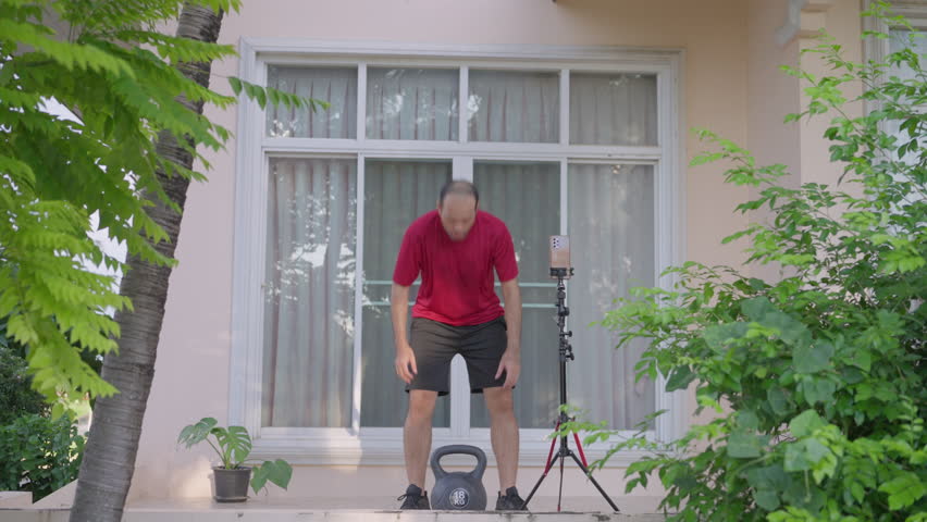 An Asian man in sports clothing exercises with a kettlebell outside his home, performing squats and recording the workout session on his smartphone to track progress and share on social media