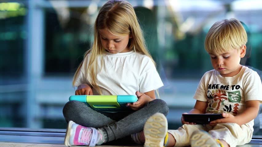Two young children sitting together using tablets, one smiling at her device, the other focused on his screen. - Powered by Shutterstock - Get 15% off with code: PIKWIZARD15