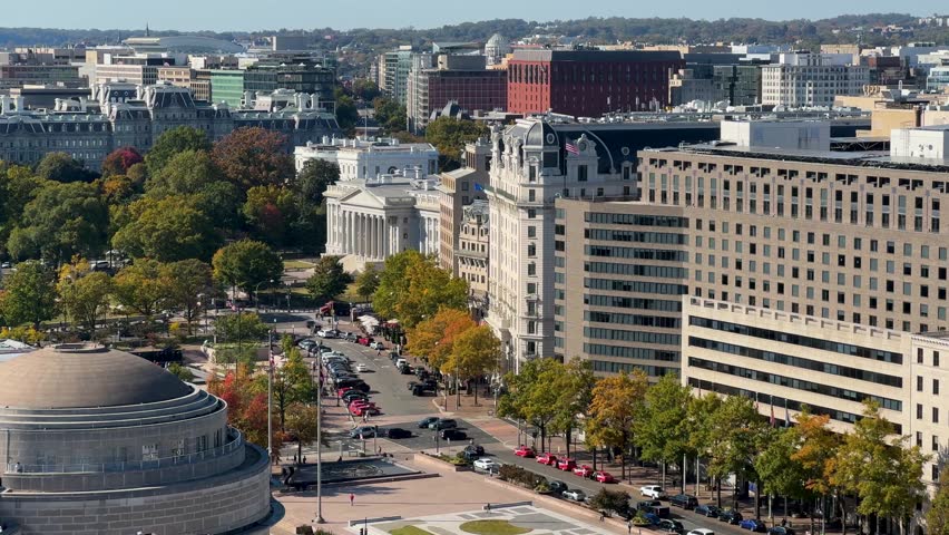A aerial view of Washington DC cityscape with White House, US Treasury, government buildings, trees, and cars. The view includes a street, and multiple trees with fall foliage.