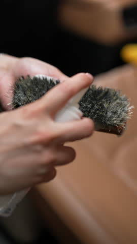 Vertical shot of unrecognizable technician male applying cleaning foam to brown leather car seat with brush, emphasizing careful attention to detail in automotive interior maintenance, slow motion.