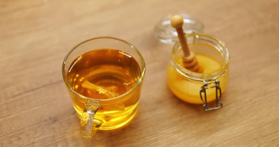 Transparent cup of aromatic tea with fresh lemon slice and honey jar stand on wooden table. Fragrant beverage brews in glass mug enriched with sour fruit