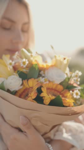 Vertical video of a blonde woman in a white dress, holding a bouquet of yellow and white flowers close to her chest. She is outside on a sunny day.
