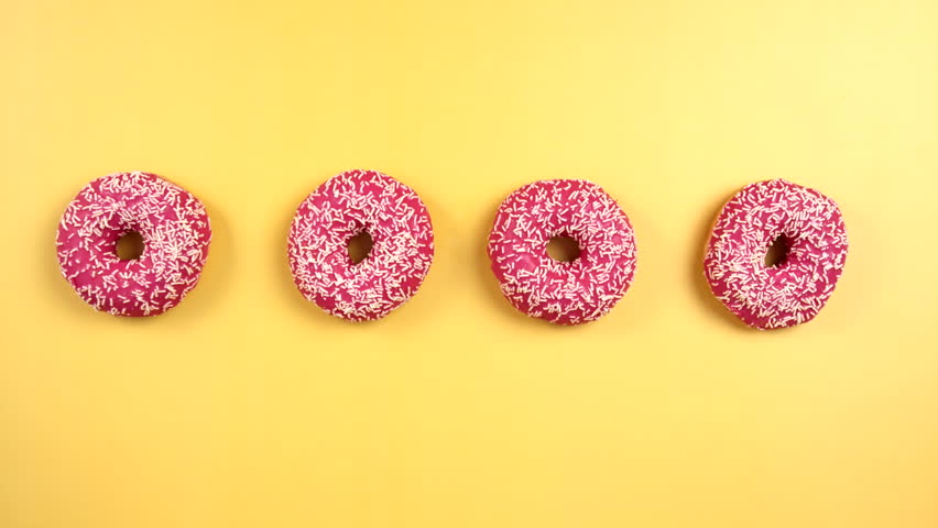 Hands grabbing four pink colorful donuts on light yellow background. Family take delicious donut at same time. Child, Male, Female. Pink glazed with sprinkles. Filmed in Prores, Topview, Slowmotion